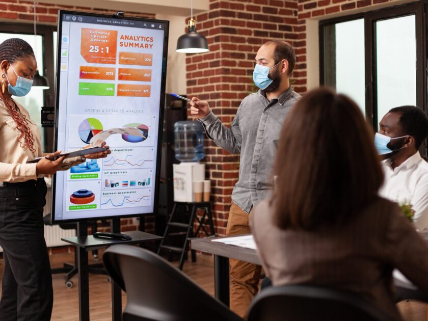 Diverse managers pointing at marketing presentation explaining financial strategy working in startup company office. Businessteam with medical face mask against covid19 brainstorming project ideas
